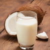 Glass of coconut milk with fresh coconut pieces on wooden surface.