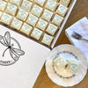 Box of white petit fours with a dragonfly logo and a plated piece beside it.