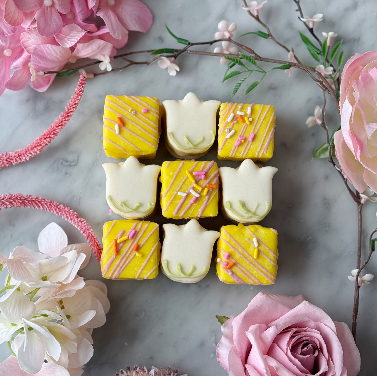 petit fours shaped like striped squares and flowers on a marble surface with pink flowers.