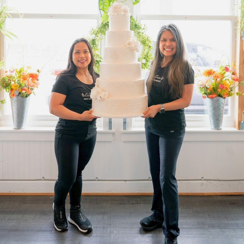 Odette D'Aniello and Mary Ann Quitugua. Two women holding a multi-tiered wedding cake indoors with flowers in the background.