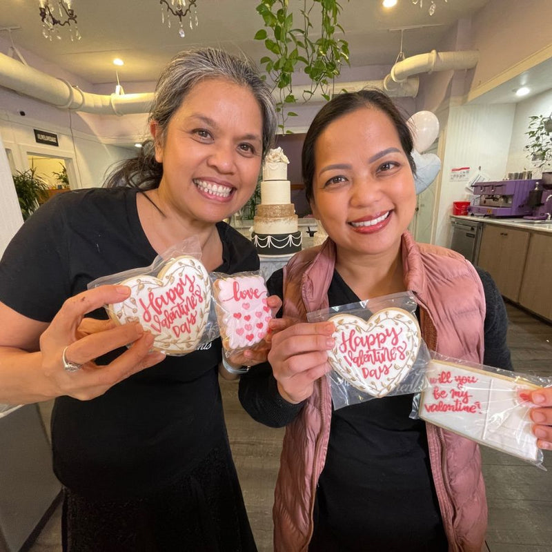 Bakery owners, Odette and Mary Ann,  holding heart-shaped Valentine's Day cookies in a bakery setting.
