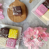 Gift set with pink floral petit fours, tea, and candles on wooden board.