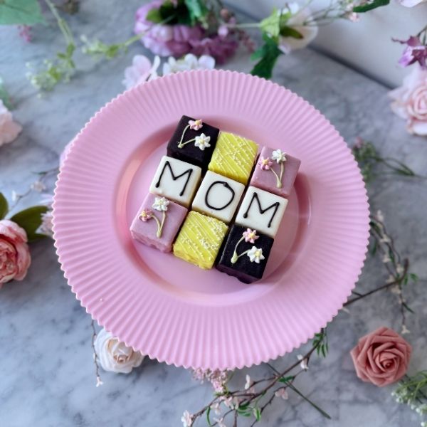 Colorful petit fours with 'MOM' decoration on a pink plate surrounded by flowers.