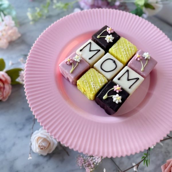 Colorful petit fours arranged to spell 'MOM' on a pink plate with flowers in the background.