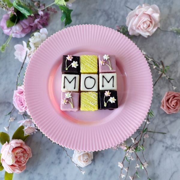 Decorative plate with 'MOM' pretit fours surrounded by flowers on a marble surface